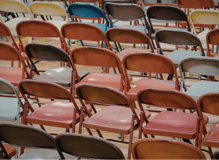 Rows of worn, multicolored metal folding chairs are arranged on a wooden floor.