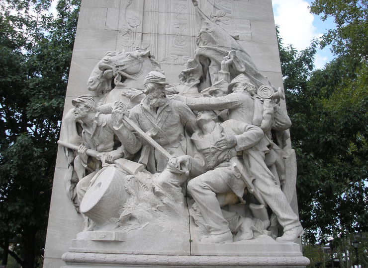 A detailed stone relief sculpture depicts soldiers, a drummer, and a flag bearer in an intense battle scene surrounded by trees.