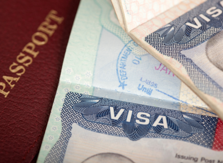 A red passport lies partially covered by two blue visa pages displaying the word “VISA” in bold white letters.