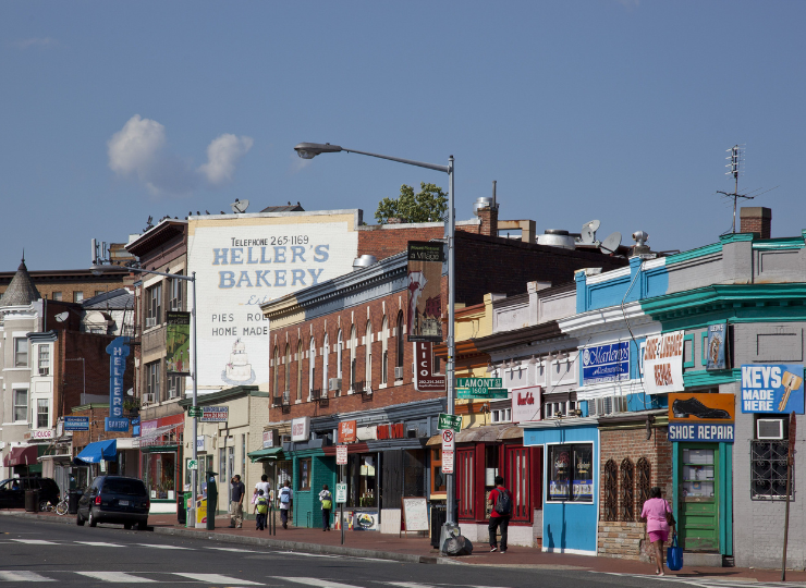 A busy neighborhood street features small local shops, vintage painted signage, and pedestrians walking past parked cars.