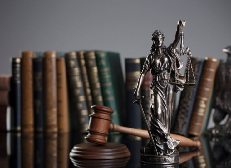 A metallic Lady Justice statue holding scales and a sword stands beside a wooden judge’s gavel in front of a row of old law books.
