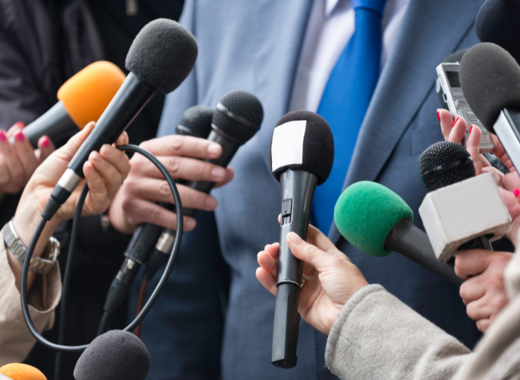 Several reporters extend colorful microphones toward a person in a blue suit during a press interview.