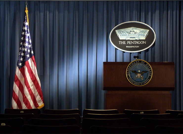A Pentagon press briefing room with an American flag, a Department of Defense podium, and rows of empty chairs in front of a blue curtain backdrop.