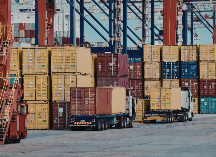 A busy shipping port filled with stacked cargo containers, trucks transporting them, and large cranes in the background.