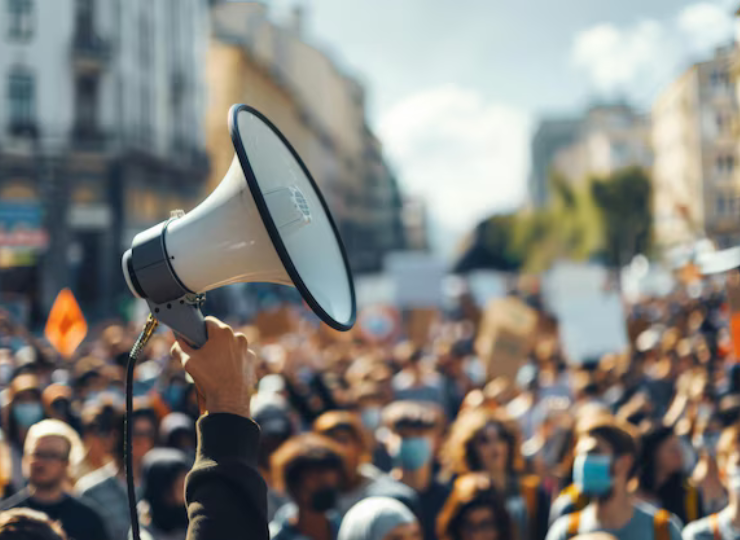 A raised hand holding a megaphone stands out above a large crowd of protesters gathered on a city street.