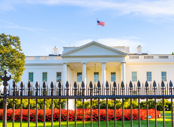 A frontal view of the White House shows its white neoclassical façade with columns, an American flag flying above the roof, and a black iron fence and landscaped lawn in the foreground.
