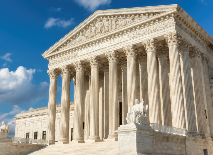 A low‑angle view of the United States Supreme Court building shows tall marble columns, a triangular pediment with carved figures, and a seated statue at the entrance beneath a bright blue sky.