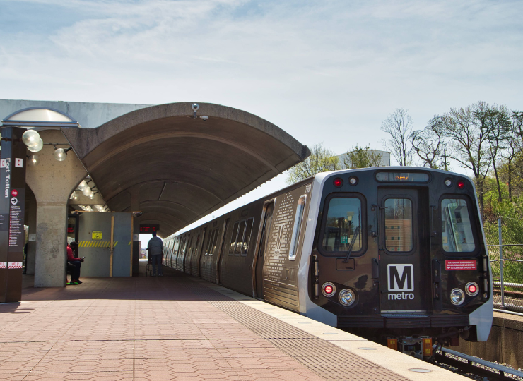 A WMATA Metro train sits at an outdoor station platform beneath a curved concrete canopy as a few passengers wait nearby.