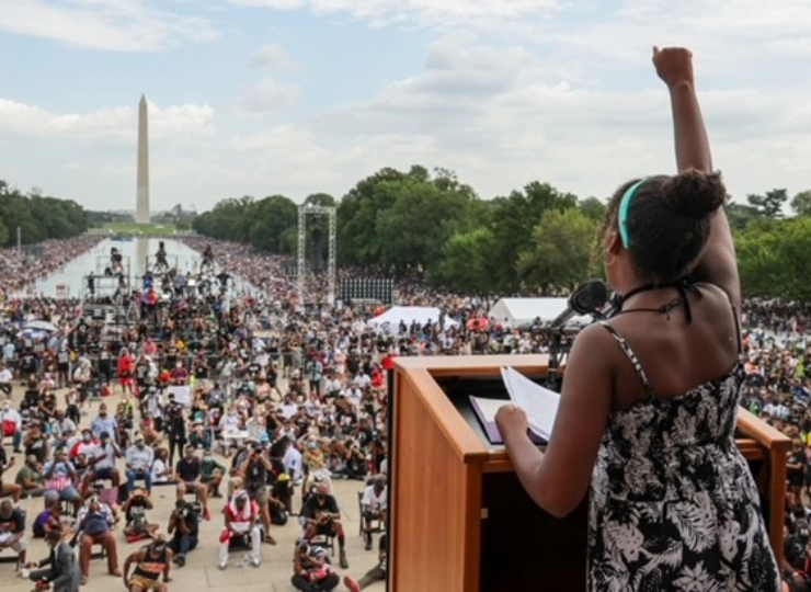 A person stands at a wooden podium with a microphone, raising one arm while facing a large crowd gathered along the National Mall in Washington, D.C. The Washington Monument is visible in the distance, with rows of seated attendees, media equipment, trees, and tents filling the open area under a partly cloudy sky.