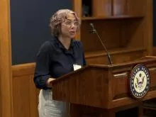 A person standing at a podium wearing a dark blue blouse and light colored pants.