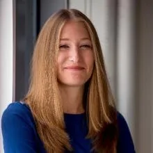 Smiling woman with straight, light brown hair wearing a blue top, standing by a window with natural light.