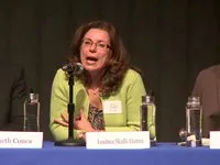 A woman wearing a light green cardigan and patterned top is seated at a panel table with a microphone in front.