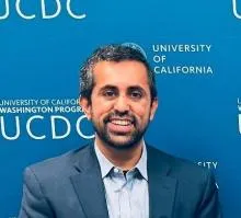 Person wearing a dark suit jacket over a light blue collared shirt, seated in front of a blue backdrop with white text that reads “UCDC.”