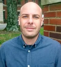 A person wearing a blue button-down shirt with the top button undone, standing outdoors near a brick wall.