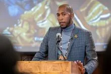 Person standing behind a wooden podium with a microphone, wearing a gray turtleneck under a textured gray blazer. 