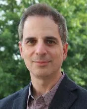 Person wearing a dark textured blazer over a patterned button-down shirt, standing outdoors with a background of lush green trees. 