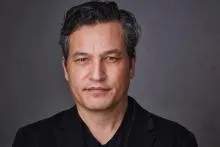 Person wearing a black blazer over a black V-neck shirt, with dark wavy hair that has gray streaks. 