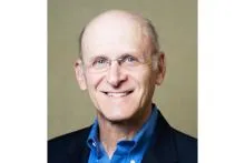 Smiling older man with glasses wearing a blue shirt and dark blazer against a neutral background.
