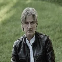 Person in leather jacket and white shirt with grass in the background.