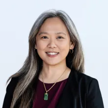 A person with long gray hair wearing a dark blazer and burgundy top poses for a professional portrait against a light background.