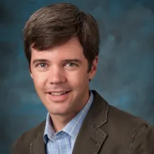 Head-and-shoulders portrait of an adult person wearing a brown jacket over a light blue collared shirt, photographed indoors against a blue studio-style background.