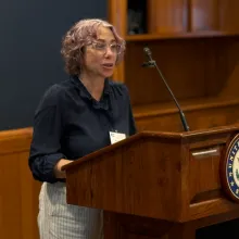 A person speaking at a wooden podium with a microphone, wearing a dark shirt and glasses, in an indoor formal setting.