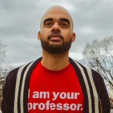 A man wearing a bright red T-shirt with bold white text that reads “I am your professor.”