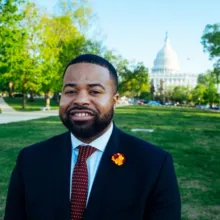 Color headshot of an adult wearing a dark suit and tie, standing outdoors on green grass with the U.S. Capitol building visible in the background.