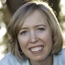 Head-and-shoulders portrait of an adult person with short light-colored hair, photographed outdoors with softly blurred foliage and light in the background.
