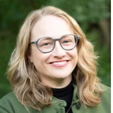 Head-and-shoulders portrait of an adult person wearing glasses and a green jacket over a dark top, photographed outdoors with foliage in the background.