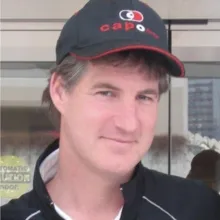 A person wearing a black baseball cap with a red‑and‑white logo and a dark jacket stands near a glass door marked with a caution sign.