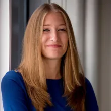 Smiling woman with straight, light brown hair wearing a blue top, standing by a window with natural light.