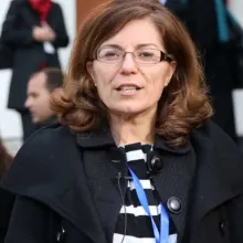 A person with shoulder‑length brown hair, glasses, and a blue lanyard stands outdoors among a crowd at what appears to be a professional event.