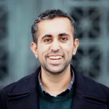 A person with short dark hair and a trimmed beard stands outdoors in front of a patterned architectural backdrop, wearing a dark coat over a green sweater and collared shirt.