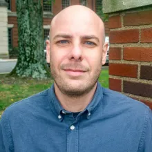 A person wearing a blue button-down shirt with the top button undone, standing outdoors near a brick wall.