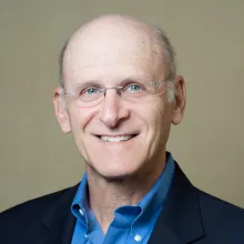 Smiling older man with glasses wearing a blue shirt and dark blazer against a neutral background.