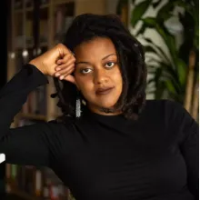 Head-and-shoulders portrait of an adult person wearing a black long-sleeve top, resting one arm against the side of the head, photographed indoors with bookshelves and plants in the background.