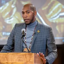 Person standing behind a wooden podium with a microphone, wearing a gray turtleneck under a textured gray blazer. 