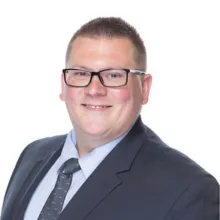 Smiling man wearing glasses, a dark suit, white shirt, and patterned tie, posing against a plain white background.
