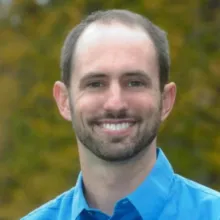 Color headshot of an adult wearing a blue collared shirt, photographed outdoors with blurred foliage in the background.