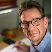 Head-and-shoulders portrait of an adult person wearing glasses and a blue collared shirt, seated at a desk indoors with papers and office items visible in the background.