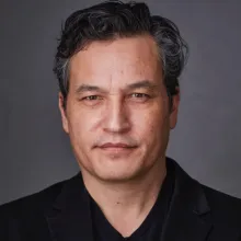 Person wearing a black blazer over a black V-neck shirt, with dark wavy hair that has gray streaks. 