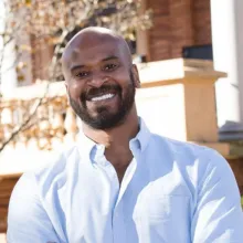 Head-and-shoulders portrait of an adult person wearing a light blue button-down shirt, photographed outdoors with a building and trees in the background.