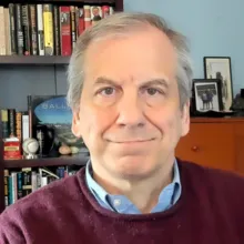 An older adult with gray hair sits indoors wearing a maroon sweater, with a bookshelf of books and personal items in the background.